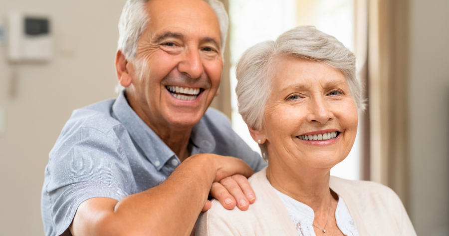 Elderly man standing behind an elderly woman, with his arms over her shoulders